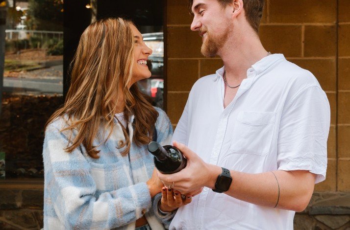 A couple looking at each other smiling and holding a bottle of wine.