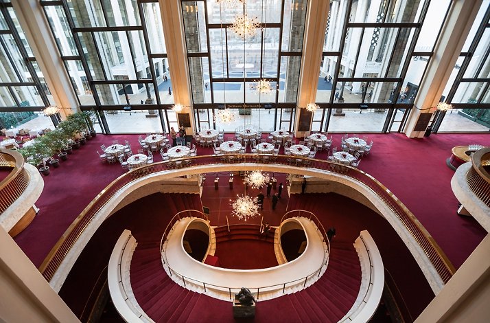 verhead view of the Met Opera’s Grand Tier lobby with sweeping curved staircase, red carpeting, and sparkling chandeliers by tall glass windows.