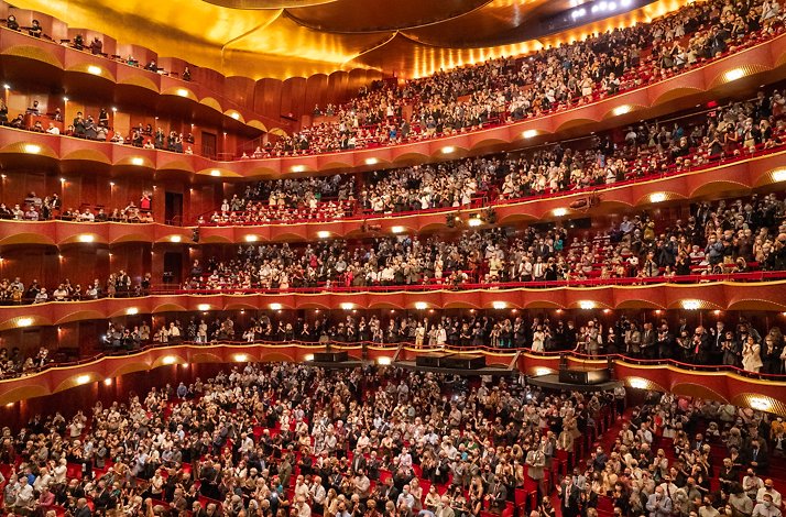 Packed The Metropolitan Opera interior with multiple red-and-gold balconies and a standing audience applauding.