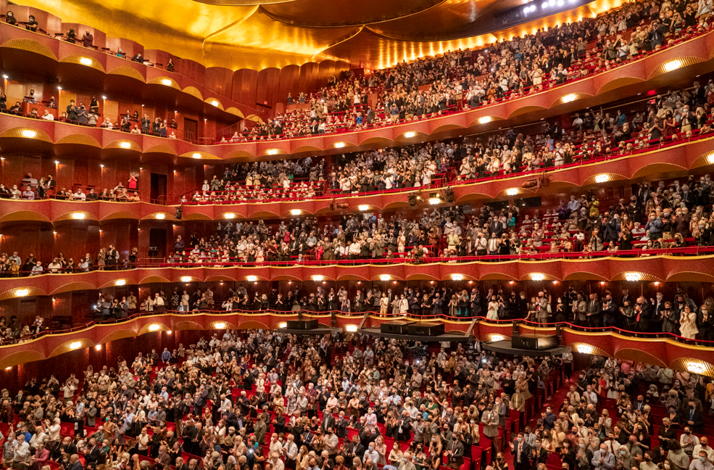 The image shows the interior of the Metropolitan Opera House in New York City, which is part of Lincoln Center for the Performing Arts with a lot of persons