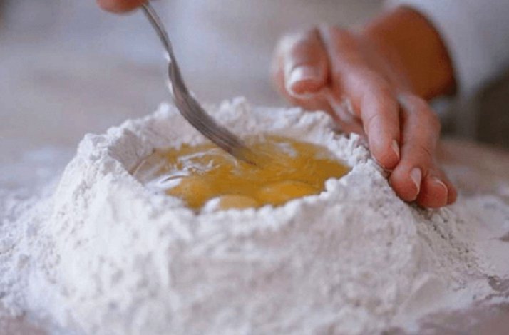Hands starting to mix a pile of flour with egg yolk with a fork.