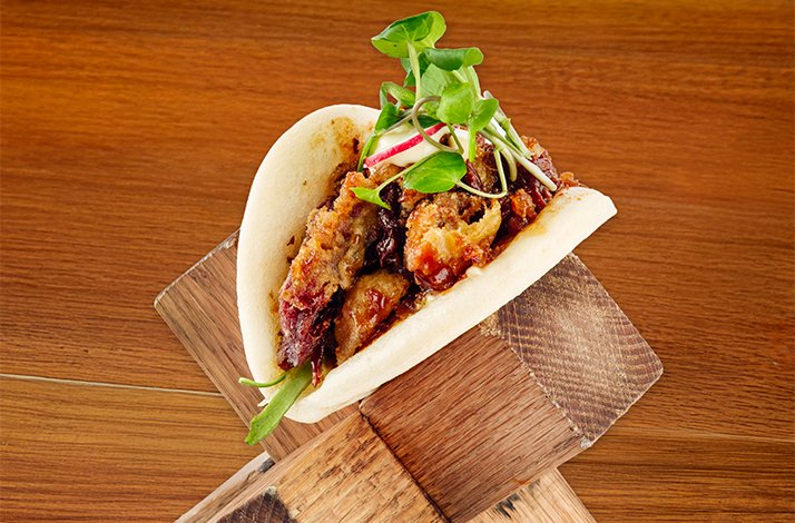 Crispy duck bao served on a wooden board, filled with glazed meat and topped with fresh microgreens and radish slices.