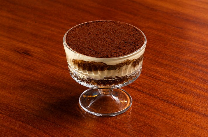 Classic tiramisu in a footed glass cup, showing distinct layers of cream, ladyfinger sponge, and a rich cocoa dusting on top.