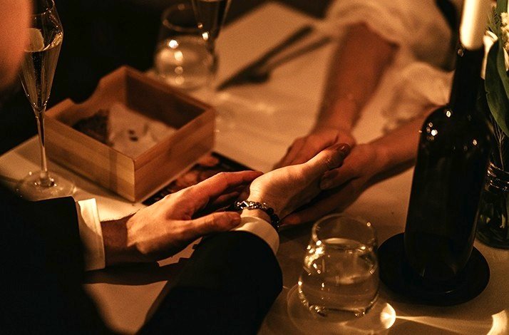 A close-up of man’s and woman’s holding hands during dinner at Da Pino. There is cutlery, wine glasses and bottle of wine on the table.
