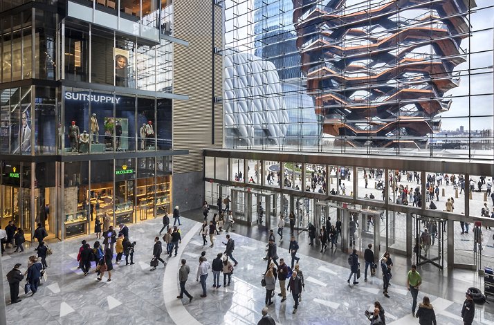A bustling retail concourse showcasing storefronts and the Vessel through glass walls.