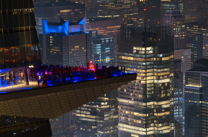 Close-up of Marquee Sky Deck, crowded with people, glowing blue lights, surrounded by skyscrapers.