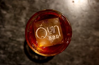 A crafted drink garnished with the Quin Bar logo.