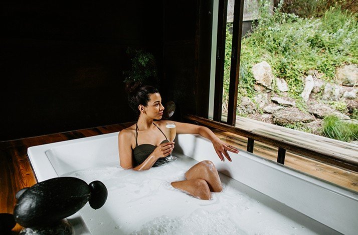 A model drinking champagne in a bath while admiring the view of nature at the Peninsula Hot Springs resort