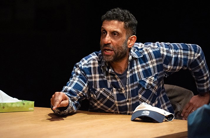 Adeel Akhtar sits at a table, gesturing during an argument.