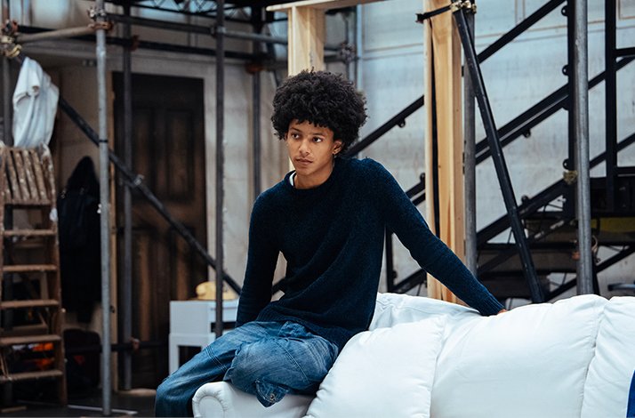 Leo Whitfield-Harris seated on a white sofa amid scaffold staging during Ilford Boy rehearsals at Donmar Warehouse. 
