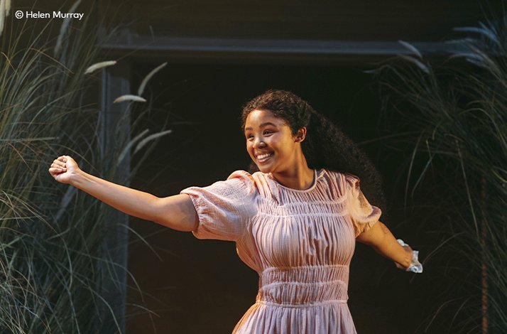 A young cast member in a pale pink dress smiling joyfully, arm outstretched, in an exuberant scene from 'A Month in the Country' at the Donmar Warehouse. © Helen Murray.