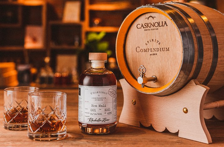 A Compendium Spirits wooden barrel, a bottle of barrel aging spirit and two glasses on a table.