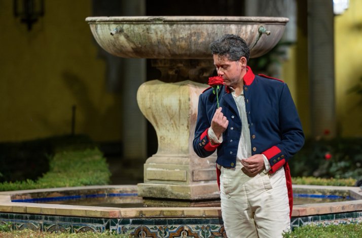 Performer in 19th-century military costume smelling a red rose beside a stone fountain at Palacio de Las Dueñas.