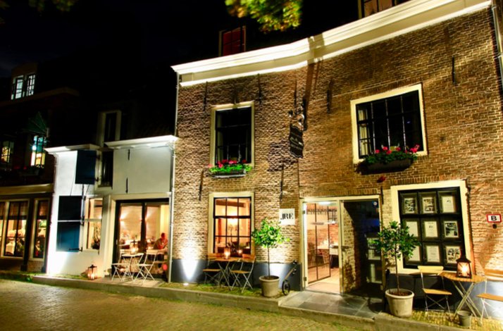 Night view of Scherp & Slapen's lit brick exterior with cafe tables, potted plants, and warm interior lighting on a quiet street.