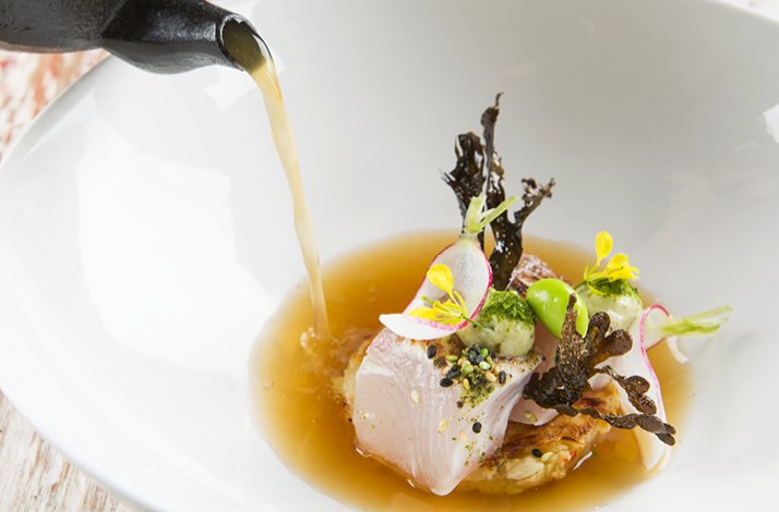 Fine dining dish receiving a golden broth pour, garnished with yellow flowers, radish slices, greens, and seaweed crisps.