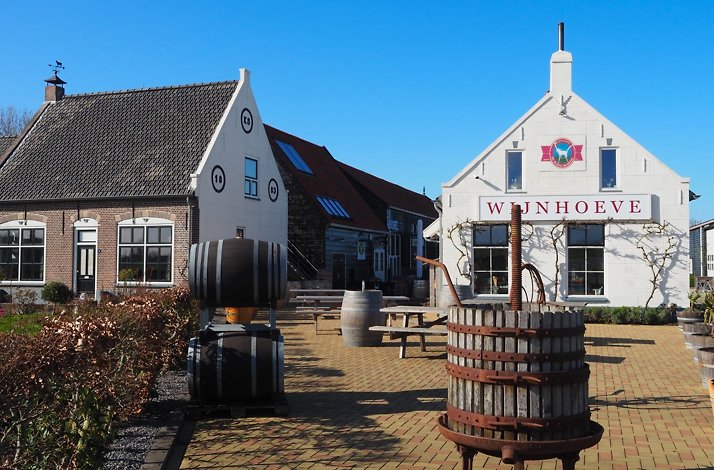 Wijnhoeve winery courtyard featuring a vintage wooden grape press, wine barrels, and traditional Dutch farm buildings under a clear blue sky.