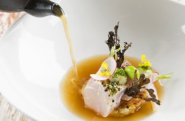 Fine dining dish receiving a golden broth pour, garnished with yellow flowers, radish slices, greens, and seaweed crisps.