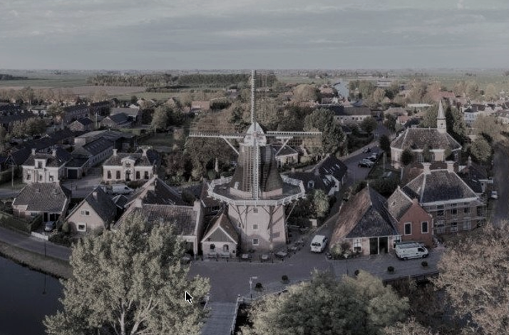 Windmill centered in a small Dutch village, surrounded by houses and trees, with canals and flat countryside stretching to horizon.