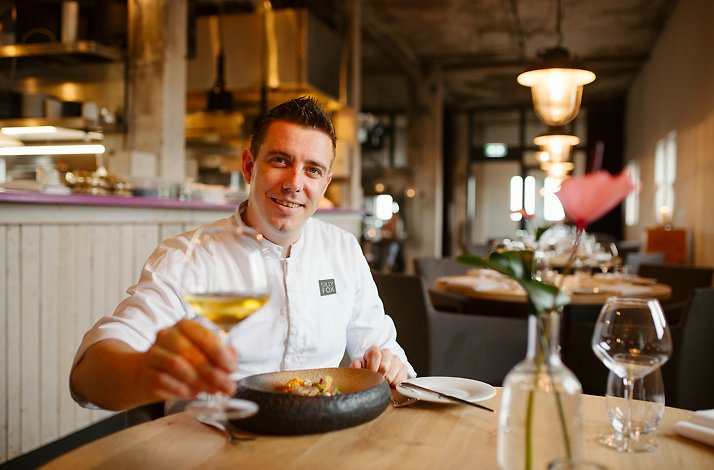 Man sitting at table in Sillyfox restaurant. Man is raising a glass of wine and has a dish on the table in front of him.