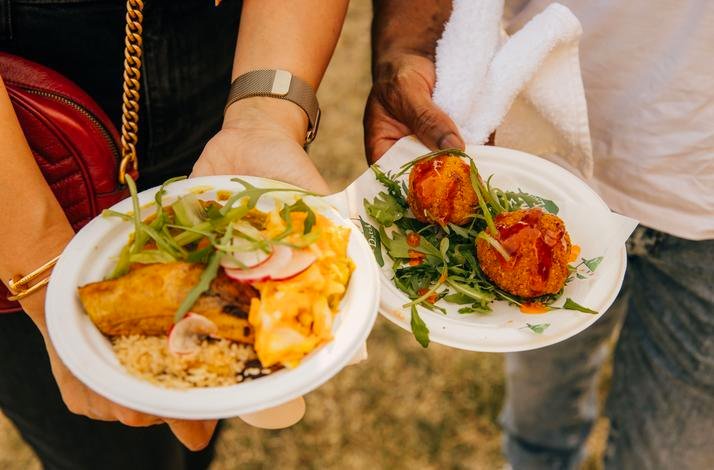 Two people holding plates of colorful festival food with arugula, rice, and fried bites at Taste of London.