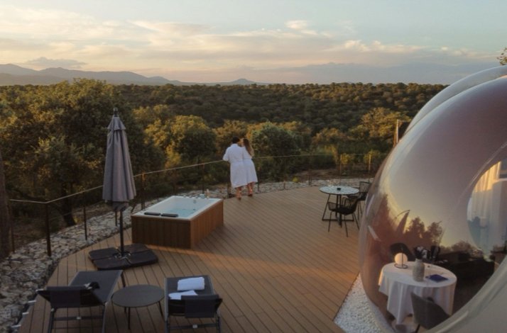 Couple in robes standing on Miluna's wooden deck at sunset, gazing over forested hills beside an outdoor hot tub and bubble suite.