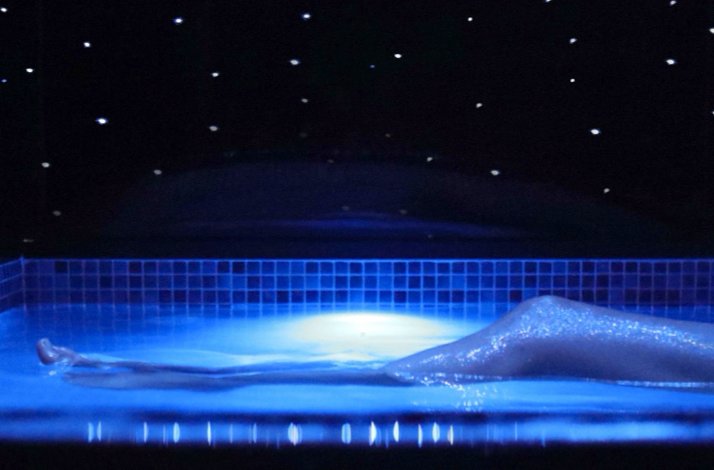 Person floating weightlessly in a blue-lit flotarium pool beneath a star-dotted ceiling during a private session.