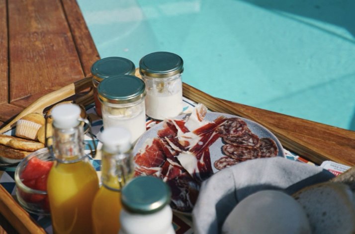 Close-up of a poolside in-room breakfast tray with Spanish charcuterie, bottled juice, and jarred condiments.