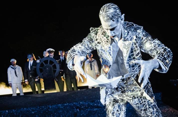 A performer in a reflective costume with sailors in the background, likely a theatrical scene.