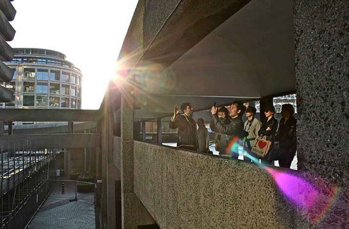 A group on a tour walking along a concrete balcony at the Barbican Estate during a sunny day.