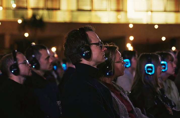 Audience wearing headphones, focused on the outdoor cinema screen at night.