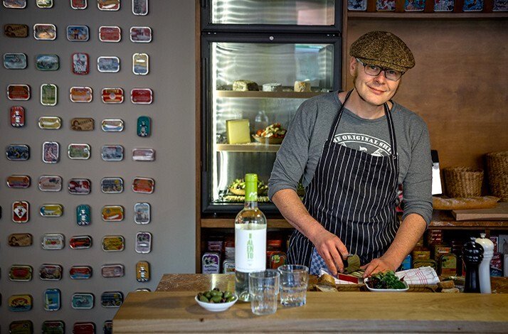 Chef in striped apron and flat cap preparing food at a wooden counter, with a wine bottle and olives in the foreground.