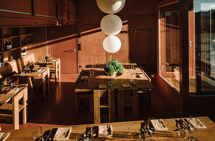 Warmly lit restaurant interior with rustic wooden tables set for dining, globe pendant lights, and a green centerpiece.