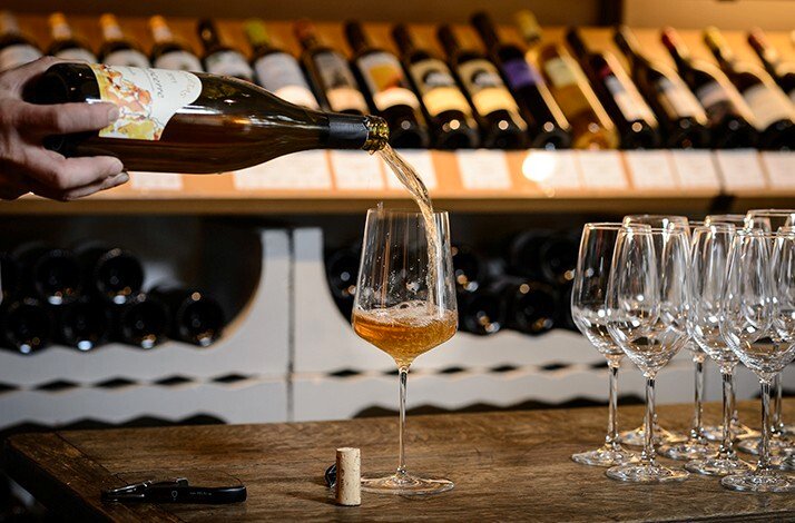 A bottle of wine being poured into a glass on a wooden counter, with several empty wine glasses and shelves of wine bottles in the background at Restaurant Brikz.