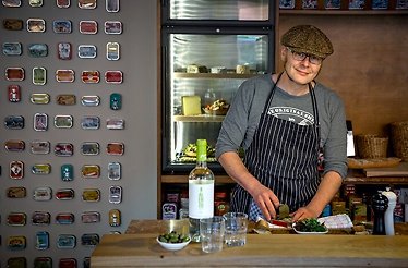 Chef in striped apron and flat cap preparing food at a wooden counter, with a wine bottle and olives in the foreground.