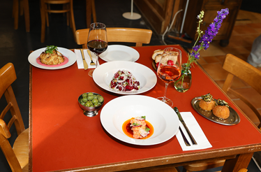 Table set for a culinary tour meal, with plated appetizers, olives, bread, and glasses of red and rosé wine beside a small vase of purple flowers.