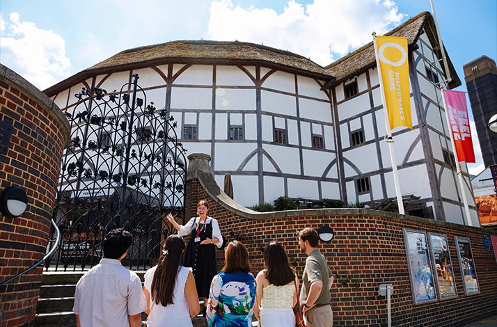 Tour guide addressing a group of five visitors outside Shakespeare's Globe Theatre on a sunny day.