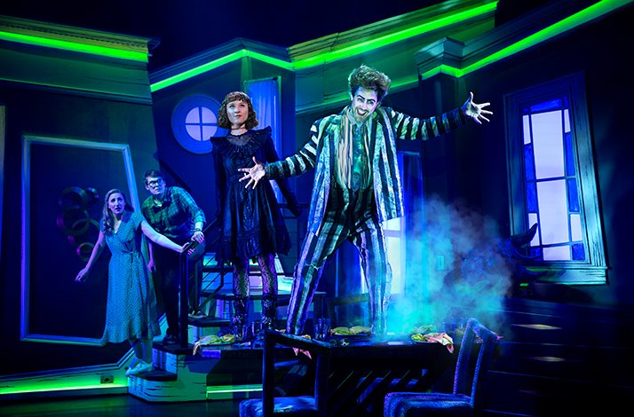 Beetlejuice stands on a table with dramatic lighting in a haunted house setting