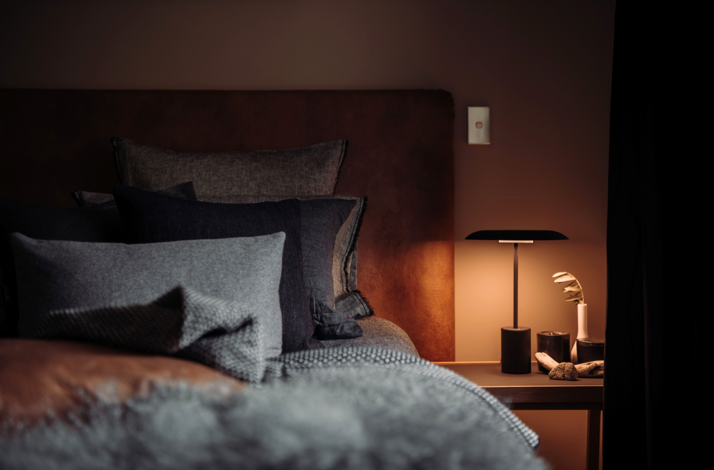 Cozy bedroom with layered pillows, warm lighting, and a bedside table.