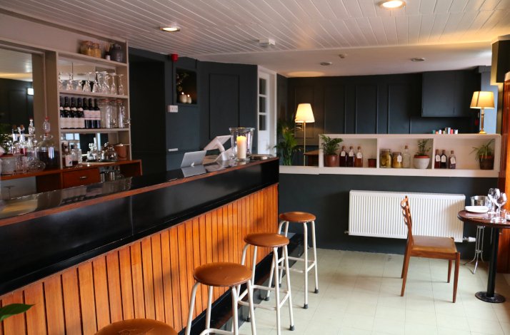 A modern bar with stools, glasses, and bottles on shelves, and a cozy seating area with a wooden chair