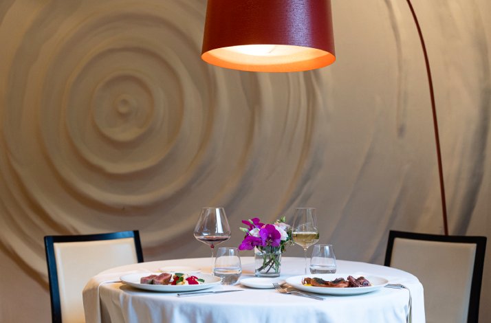 Intimately set table for two with plated courses, stemware, and purple blooms beneath a red arc lamp at Dubravkin put.