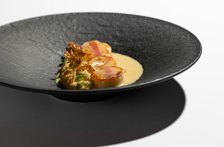 Seared scallops with bacon and golden sauce, plated on a dark matte dish, as part of Dubravkin put's tasting menu.