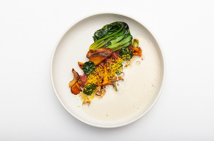 Fine-dining course at La Leggenda dei Frati presenting wilted bok choy, chanterelles, golden crumble, and roasted vegetables on white sauce.