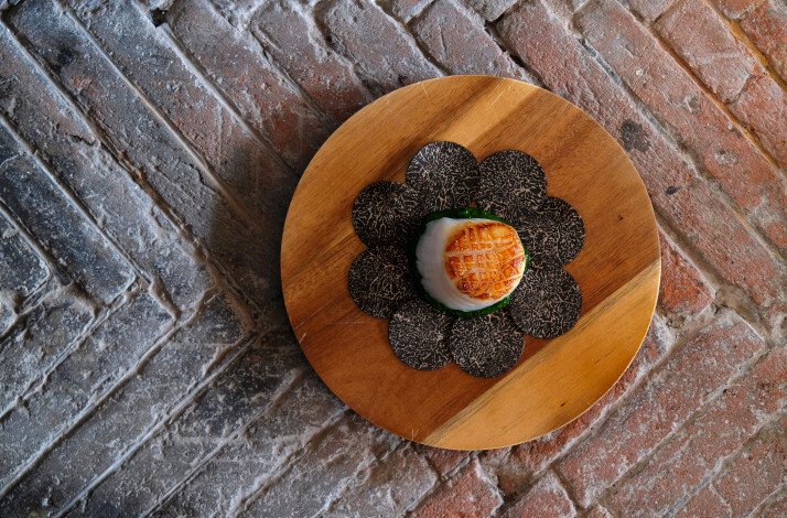 A round wooden plate with a scallop sitting in the middle and black truffle slices arranged around it served at Restaurant G.a.