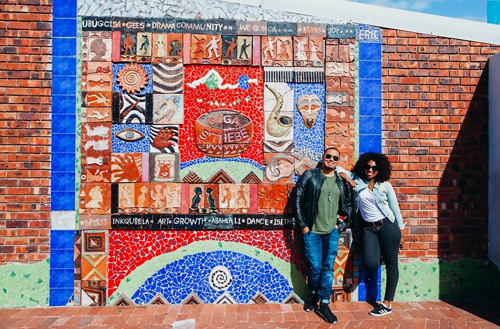 Couple standing against the colourful Guga S'Thebe cultural mural in Langa Township, Cape Town, with mosaic tiles and text.