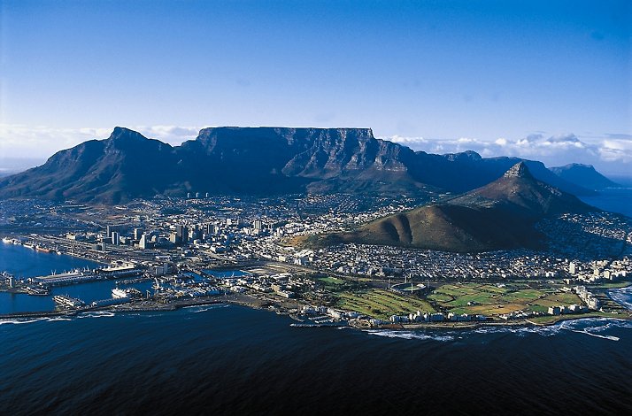 Cape Town cityscape and waterfront seen from above, framed by Table Mountain's flat summit and surrounding peaks.