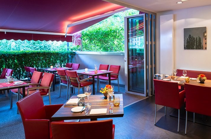 Sun-lit terrace interior at Huber Munich featuring red upholstered seating, neatly set tables, and open views of surrounding greenery.