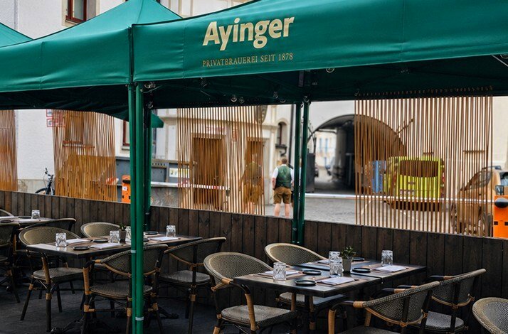 Ayinger Privatbrauerei seit 1878 branded green canopy over a set outdoor terrace with wicker chairs and wooden privacy screens in Munich.