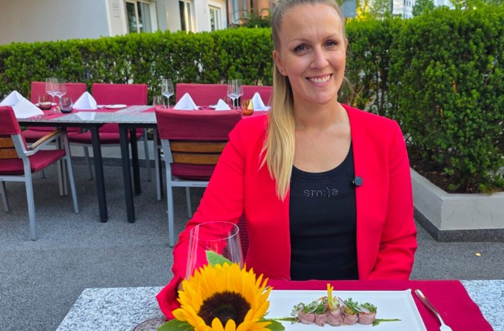 Food blogger Bianca Murthy seated at an outdoor table at Restaurant Huber, smiling in a red blazer, with a sunflower and plated dish in front of her.