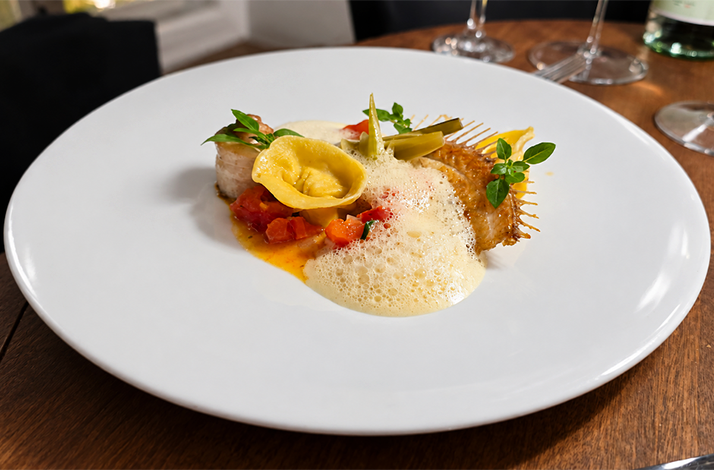 Plated fish course with foam sauce, ravioli, and vegetables on a white plate at Atlantik Fisch.