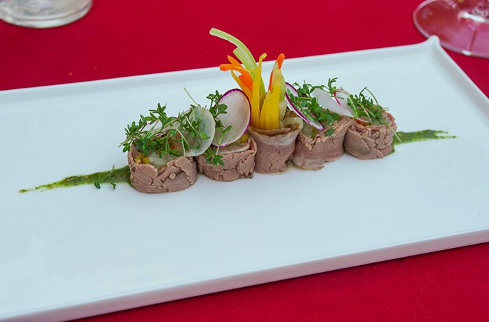 Artfully arranged meat roll appetizers topped with fresh microgreens and radish, served on a white plate with green herb sauce.